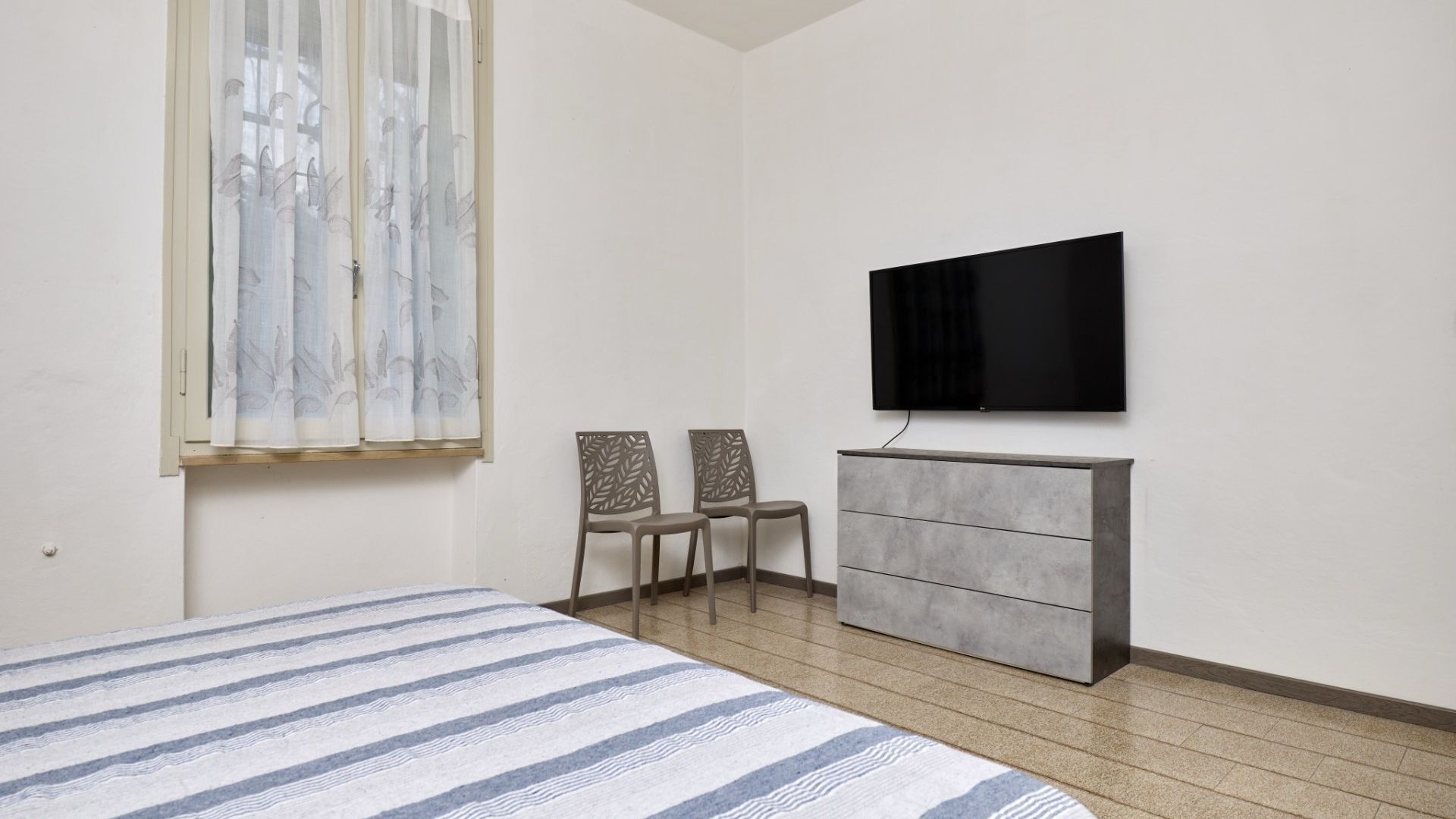 A bedroom with a bed, a dresser, two chairs, a television, and a window with sheer curtains.