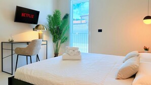 Quarto de hotel com uma cama, uma escrivaninha com cadeira, uma TV exibindo 'NETFLIX', um vaso de planta e vista para o exterior através de uma porta.