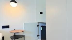 Una habitación compacta con una cama, una mesita redonda de madera, una silla negra, un mueble negro con un refrigerador en su interior y una lámpara colgante.
