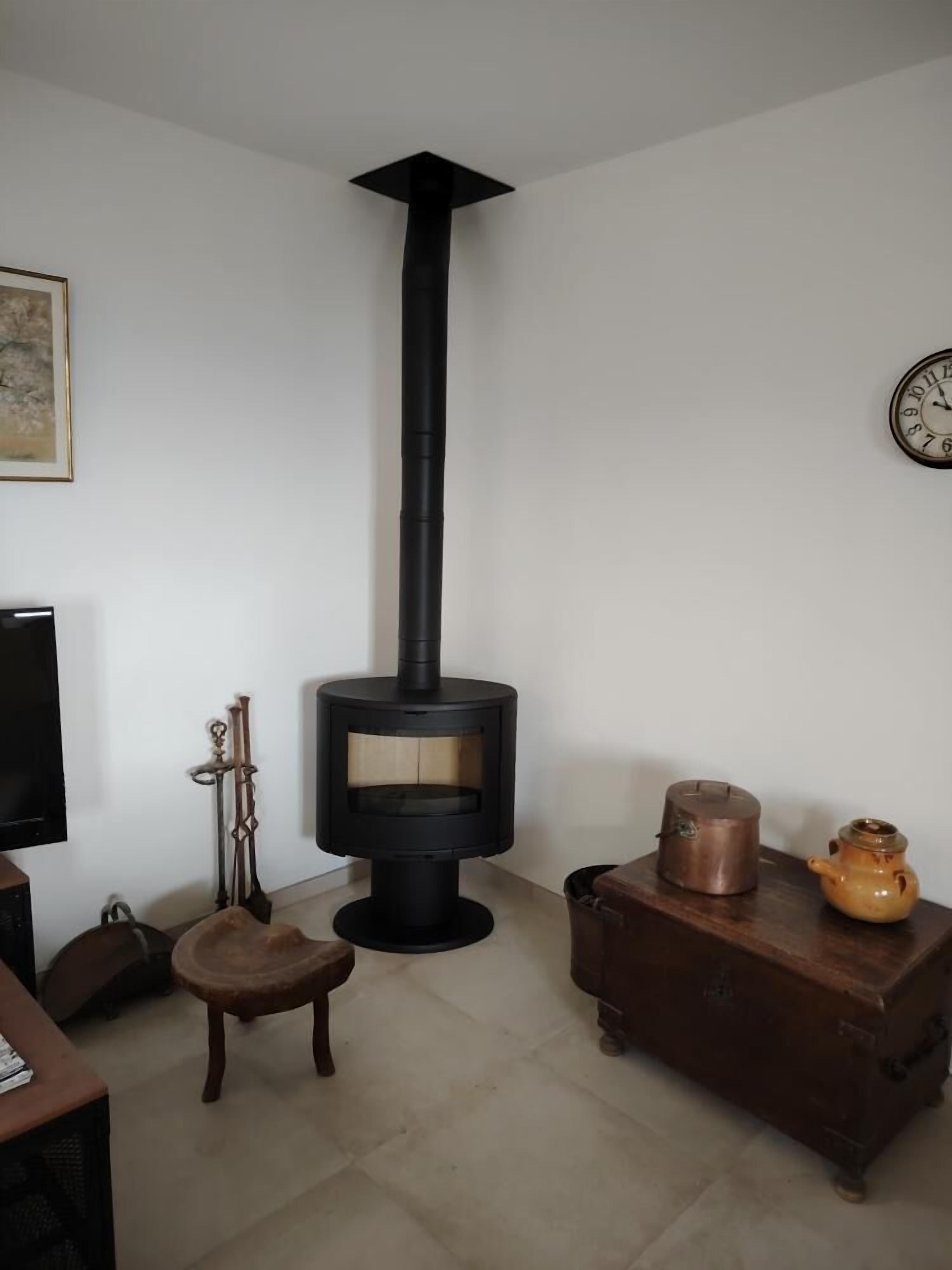 Una habitación con una estufa de leña negra, una silla de madera, una mesita con un calentador y un reloj en la pared.