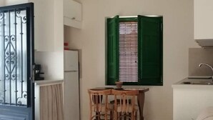 A compact kitchen with a dining area, a window with green shutters, and a white refrigerator. - Lacasadesowsam (Granada)