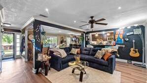 A spacious living room with a dark blue sofa, a glass coffee table, and a ceiling fan. - 6mi from AT&T-HEATED POOL-HOT TUB-INDOOR BAR-TOP KITCHEN AMENITIES-SLEEPS 12+ (Arlington)