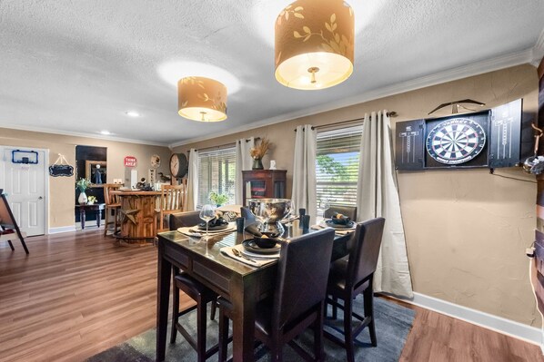Een eetgedeelte met een houten tafel voor vier personen, een dartbord aan de muur, en een keuken op de achtergrond.