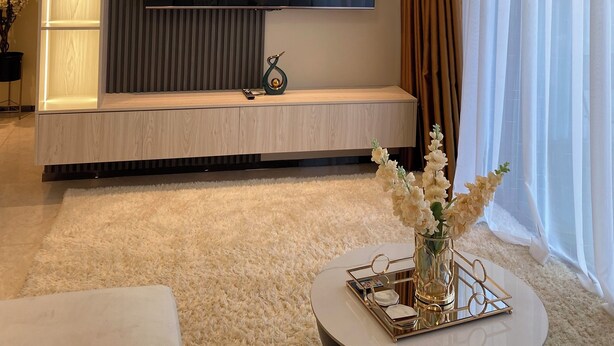 A modern living room with a flat-screen TV mounted on a wooden panel wall, a round coffee table with a vase of flowers, and a large window with sheer curtains. - Chic 1-bedroom apartment in wonderful Nairobi with AC, fitness room, WiFi (Nairobi)