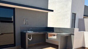 Une cuisine extérieure moderne, dotée d’un évier et d’un barbecue intégrés, située devant un bâtiment contemporain avec un toit plat et de grandes fenêtres.