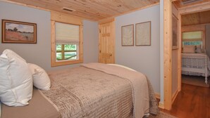 Un dormitorio con techo de madera, una cama con colcha color crema, una ventana con vistas a zonas verdes y una puerta que da a otra habitación.