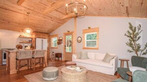 Interior acogedor de una cabaña con techo de madera, comedor con cocina, sala de estar con sofá y sillones, y un ventanal grande.