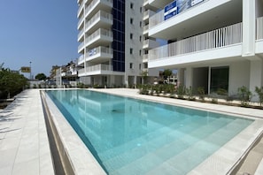 A modern hotel with a swimming pool, multiple balconies, and a clear sky.