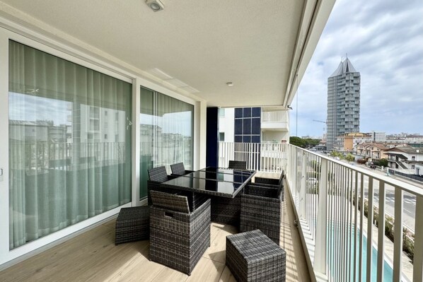 A balcony with a glass table and wicker furniture, overlooking a cityscape with modern buildings.