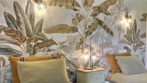 A bedroom with a mural of tropical foliage, a bedside table with a lamp, and two beds with pillows. - Des Puits - LOCOAL-MENDON (LOCOAL-MENDON)