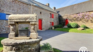 A stone well with a metal basin in front of a stone building with red doors and windows. - Des Puits - LOCOAL-MENDON (LOCOAL-MENDON)