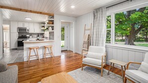 A modern kitchen with white cabinets, a central island with stools, and a view of a tree through large windows. - Remodeled 2-bedroom house with hot tub, 7mins to Saugatuck & beach (Fennville)