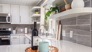 A modern kitchen with white cabinets, a stainless steel microwave, and a wine bottle on a wooden tray. - Remodeled 2-bedroom house with hot tub, 7mins to Saugatuck & beach (Fennville)