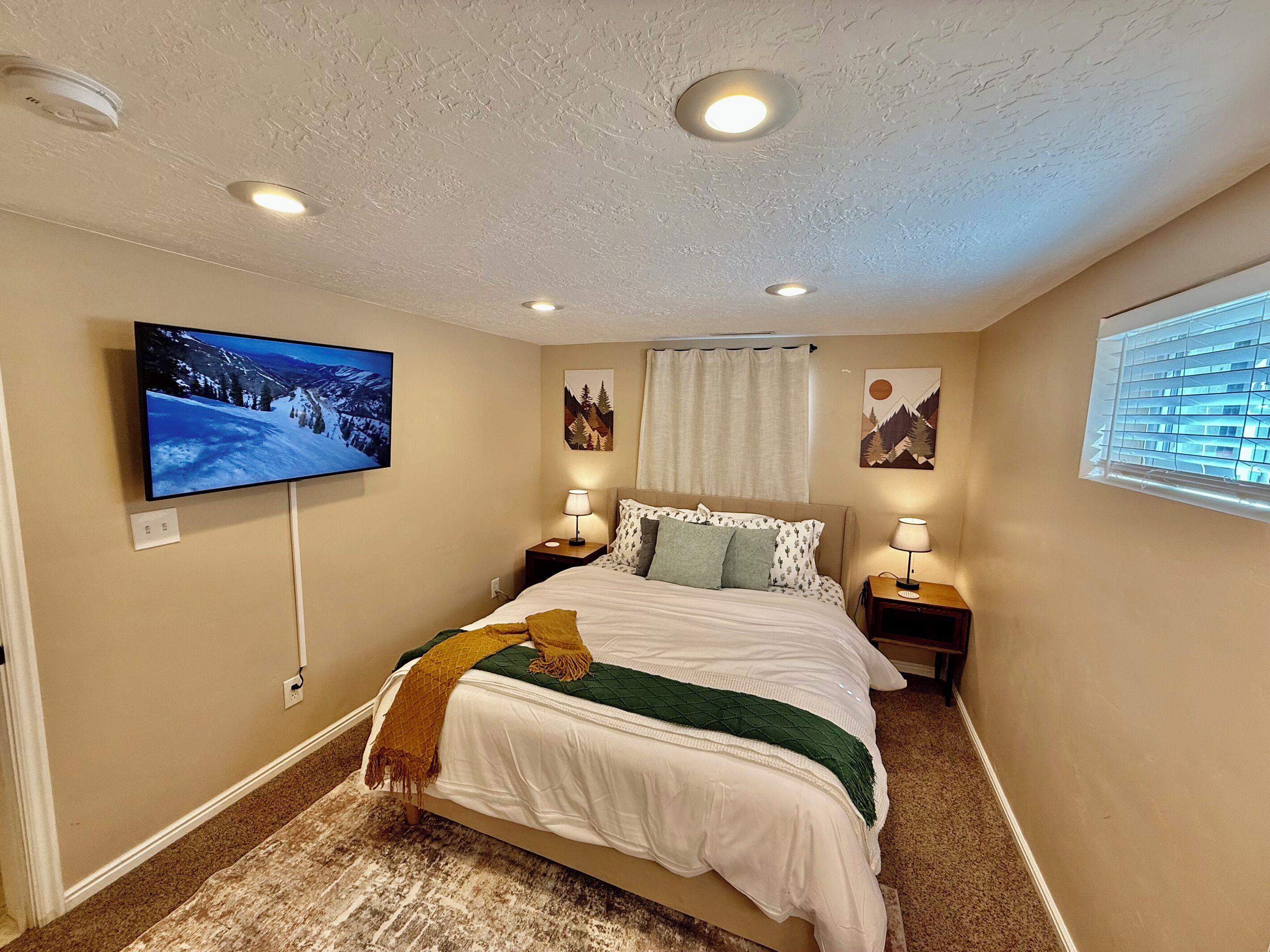 A bedroom with a bed, two bedside tables with lamps, a flat-screen TV mounted on the wall, and a window with blinds.