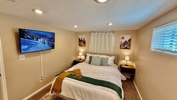 A bedroom with a bed, two bedside tables with lamps, a flat-screen TV mounted on the wall, and a window with blinds.