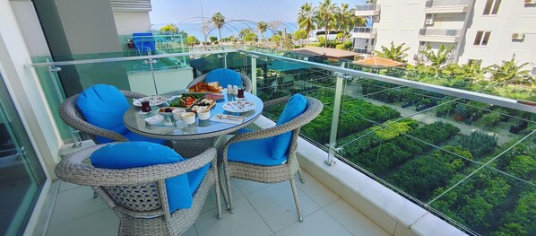Un balcón con una mesa de vidrio y sillas de mimbre, con vista a un jardín exuberante y edificios residenciales.
