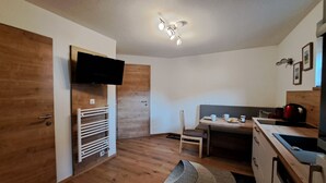A compact kitchen with wooden flooring, a built-in TV, a dining area with a bench, and a small refrigerator.