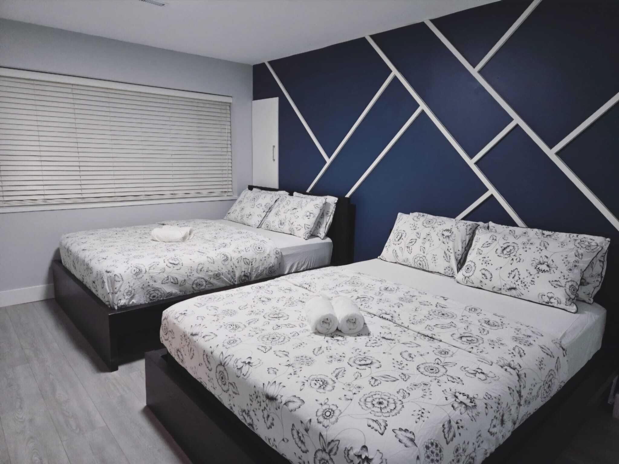 A bedroom with two beds, a window with blinds, and a geometric wall design.