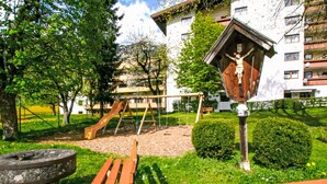 Un terrain de jeux avec des balançoires et un toboggan, entouré de verdure et bordé d’un bâtiment à la structure en bois.