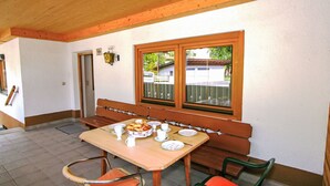 Un coin repas avec une table en bois dressée pour deux personnes, un banc et une fenêtre donnant sur une voiture garée et sur la verdure à l’extérieur.