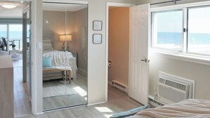 A bedroom with a bed, a window with a view, a door leading to another room, and a mirror. - BeachBox III • Unreal Views • Oceanfront Balcony • Couple's Getaway (Ocean City)