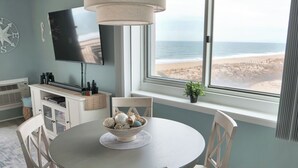 Un comedor con una mesa redonda, sillas blancas, un cuenco con adornos, un televisor en la pared y vistas a la playa a través de un ventanal grande.