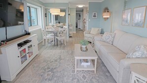 A spacious living room with a large TV, a dining area with white furniture, and a light blue wall with a 'BEACH' sign. - BeachBox III • Unreal Views • Oceanfront Balcony • Couple's Getaway (Ocean City)