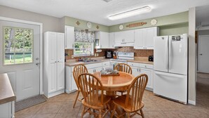 A kitchen with white cabinets, a dining table with chairs, a refrigerator, and a window with a view of greenery. - Quiet Getaway Near Ft. Myers • Relax & Unwind (Alva)
