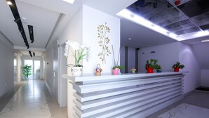 Egy modern recepciós pult, fehér és szürke csíkos mintával, melyet virágtartókban elhelyezett növények és egy díszes falfelület vesz körül.
