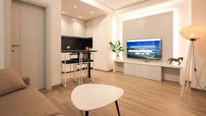 Egy modern nappali, amelyben található egy kanapé, egy étkezőasztal, egy televízió, amelyen a „GRANDSTAY APARTMENTS” felirat látható, és egy konyhasarok.