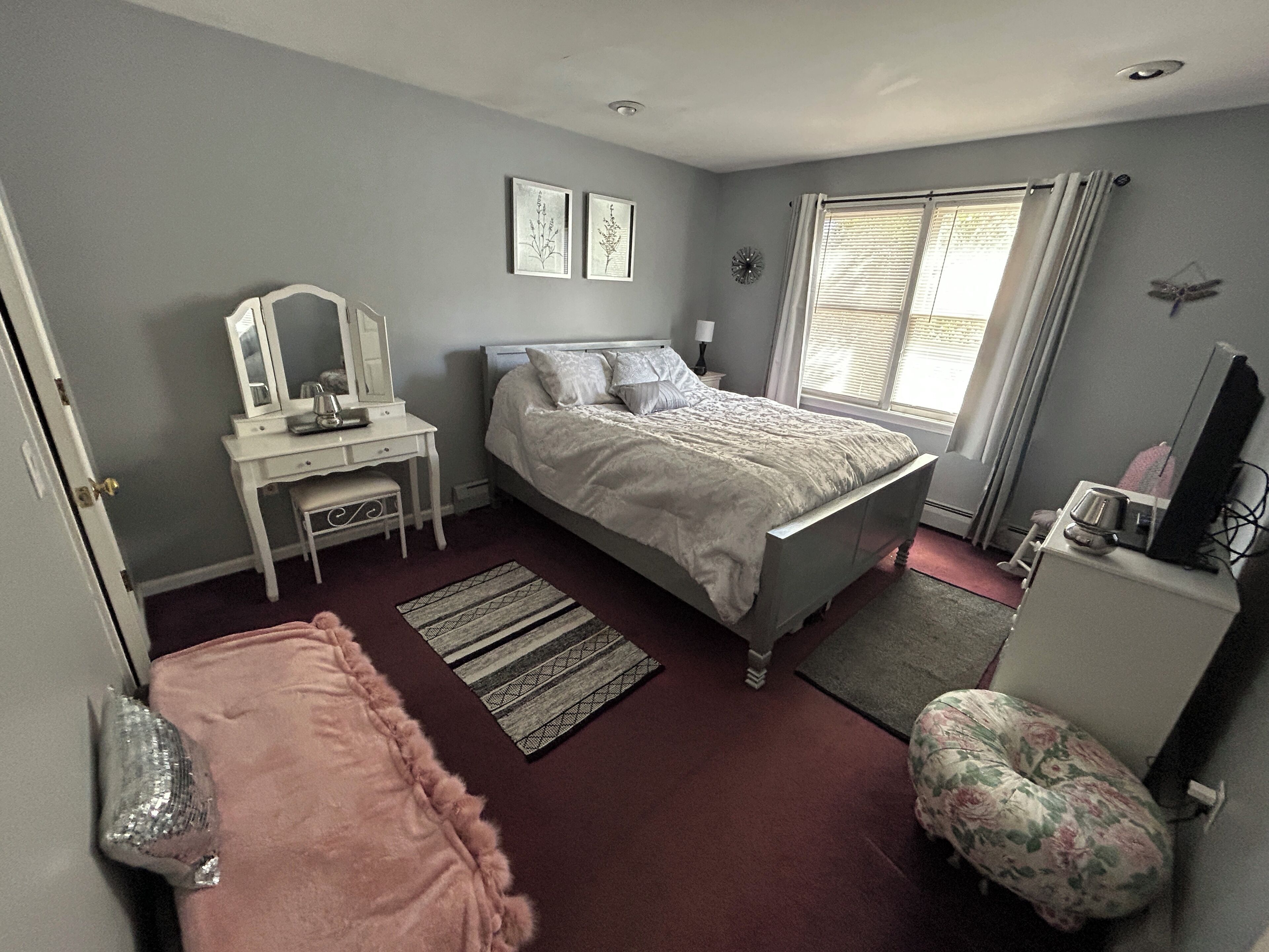 A bedroom with a bed, a dresser, a vanity with a mirror, and a window with curtains.