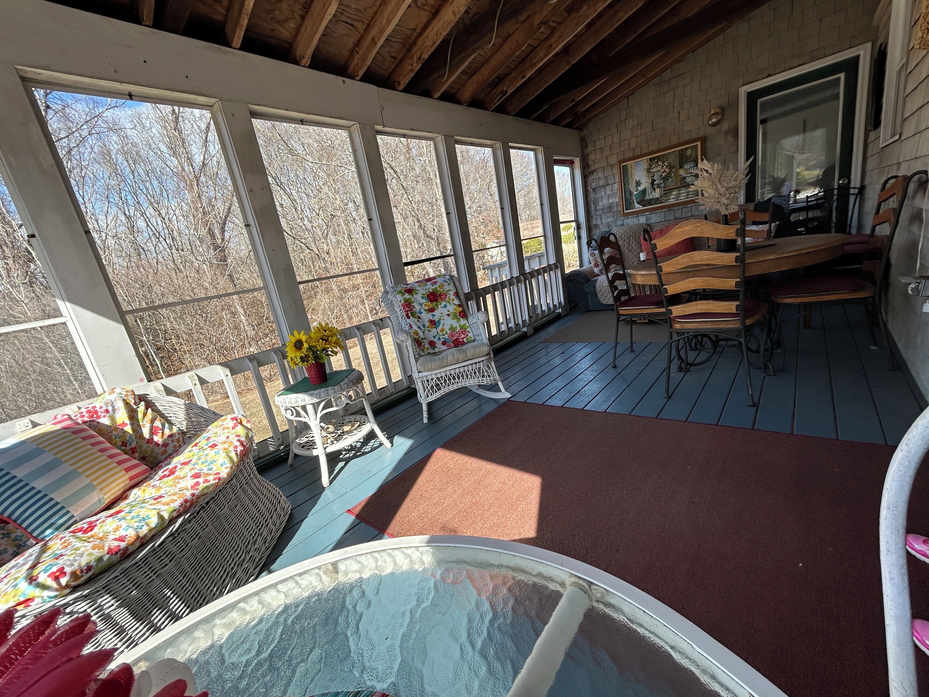 A covered porch with wicker furniture, a glass table, and a view of trees.
