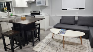 Une cuisine moderne avec des armoires blanches, un four à micro-ondes et une cuisinière. On y trouve un coin repas avec une table et des tabourets en bois foncé. Un canapé gris et une table basse blanche, sur laquelle sont posés des magazines, se trouvent dans l’espace salon.