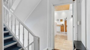 Un escalier avec des balustrades blanches mène à une pièce dotée d’un plafond en bois et dont on aperçoit une cuisine à travers une porte ouverte.