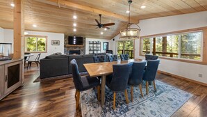 A spacious living area with a wooden ceiling, a dining table with chairs, a fireplace, and large windows offering a view of trees.