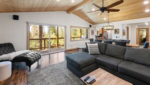 A spacious living room with a large gray sectional sofa, a black tufted ottoman, and a wooden coffee table. There is a ceiling fan and recessed lighting.