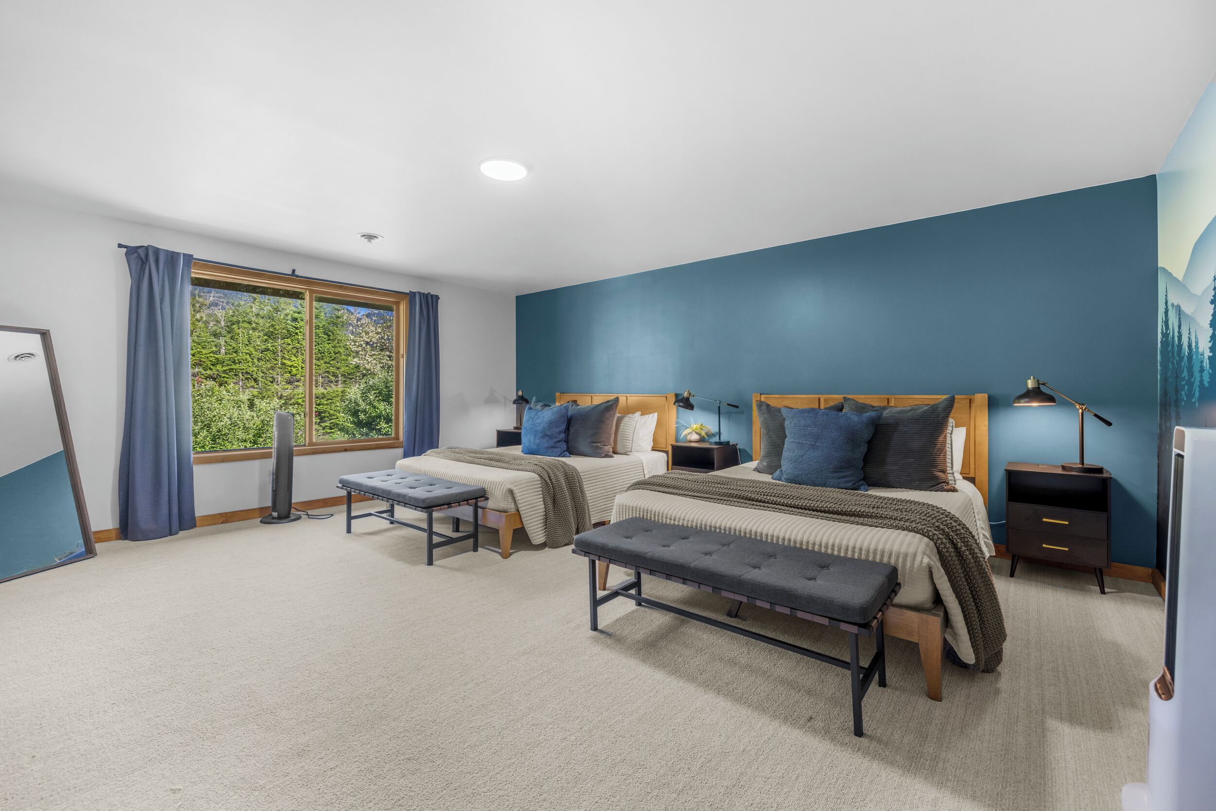 A bedroom with two beds, a window with curtains, a standing fan, and a nightstand with a lamp.