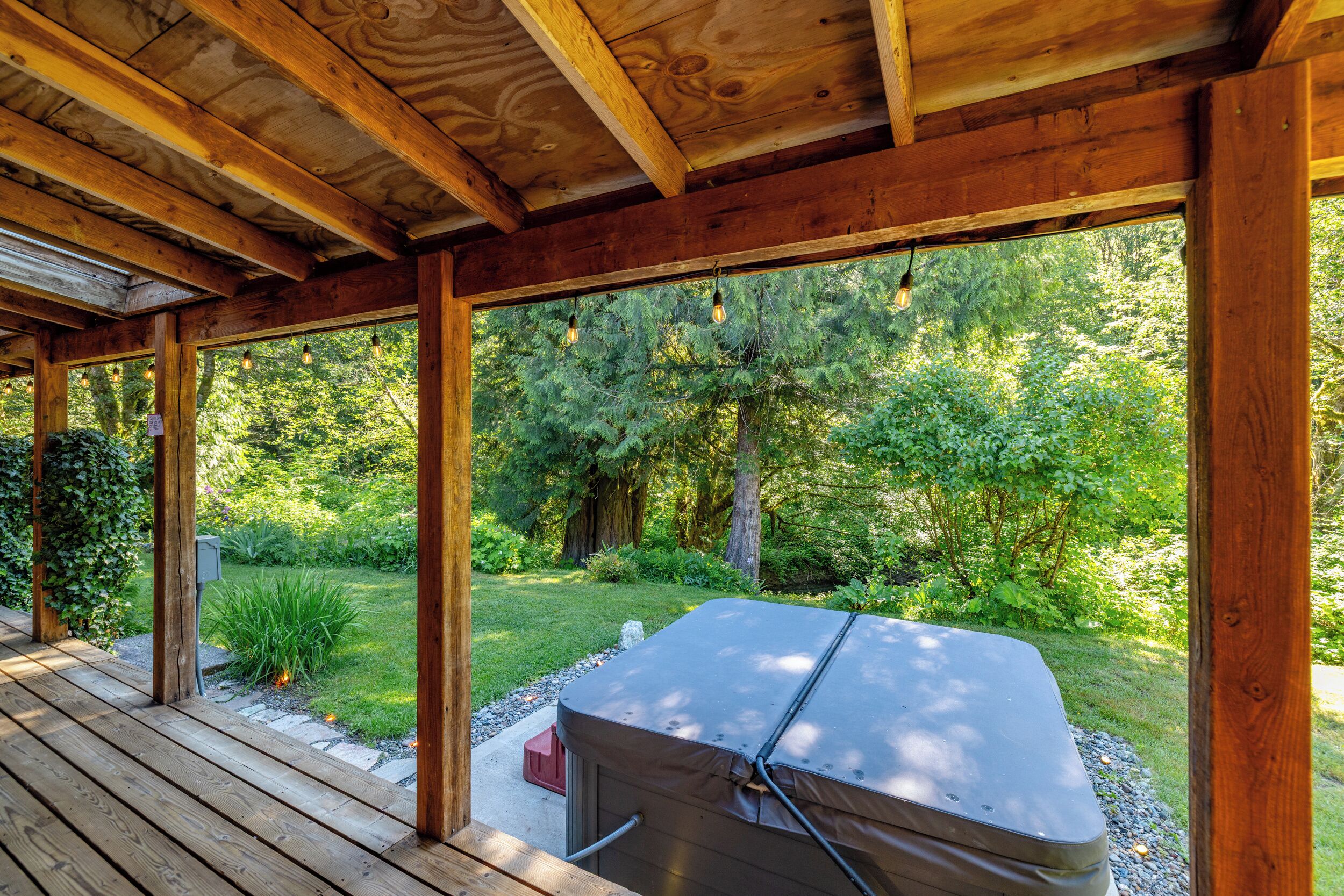 A covered outdoor area with a barbecue grill, wooden beams, and a view of a lush garden.