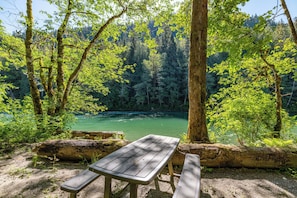 A picnic table and benches in a forested area with a river in the background. - American Alps Lodge: Riverfront, Beach, Hot Tub, EV Charger, Game Room (Marblemount)