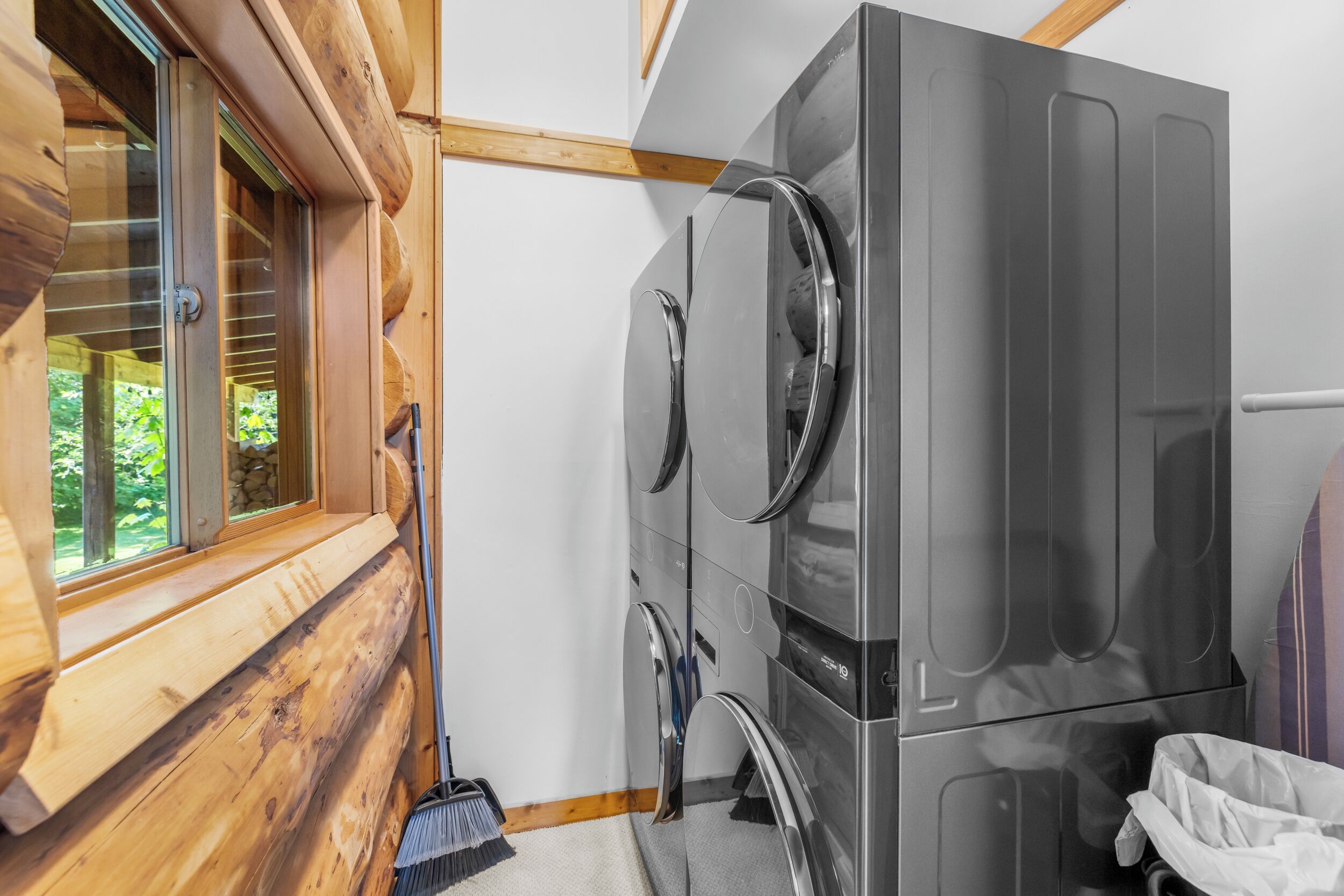 Une petite buanderie équipée d’un grand lave-linge et d’un sèche-linge en acier inoxydable, avec des murs en bois et une fenêtre dotée de stores.