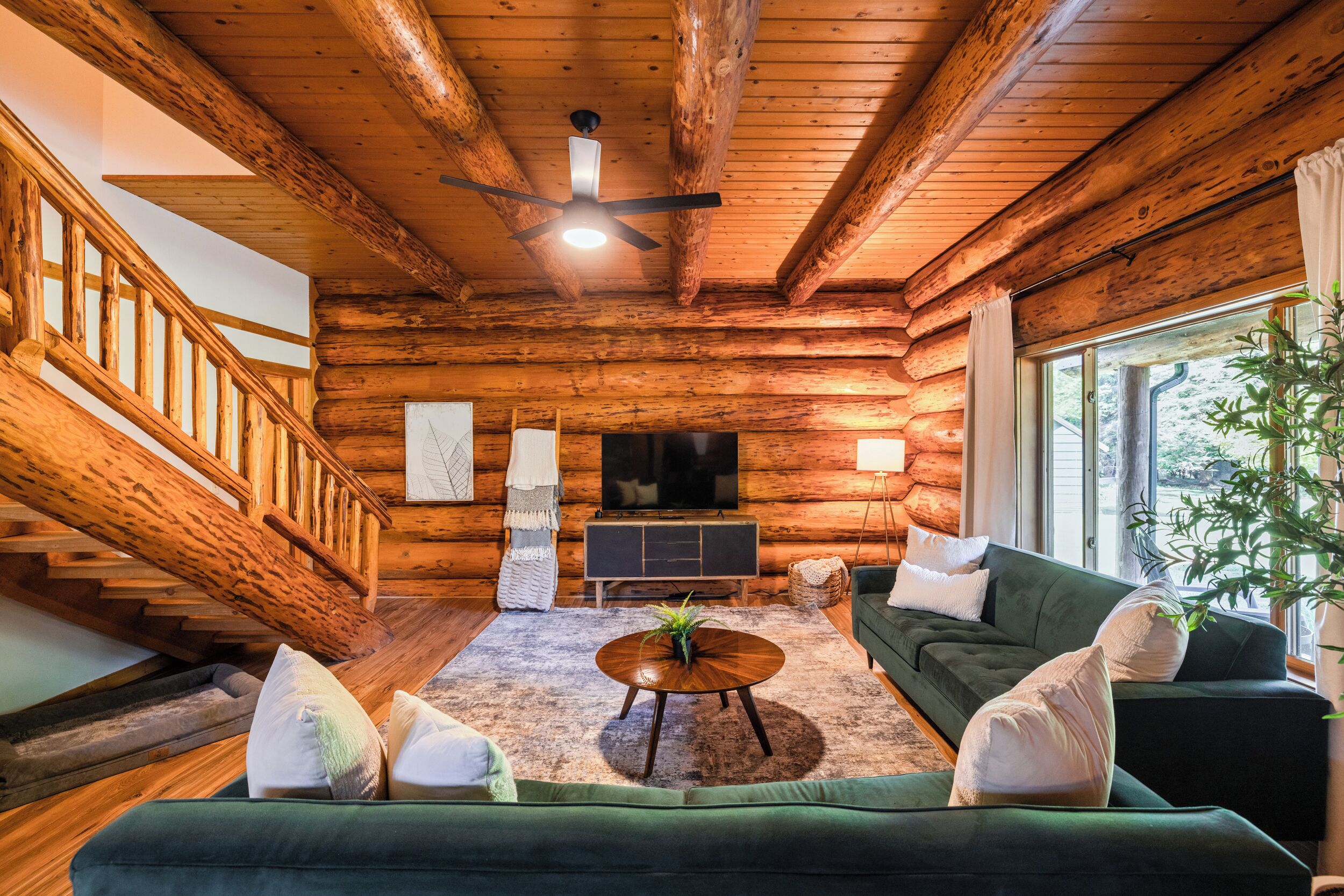 Interior de una cabaña de troncos con una escalera, un televisor, un sofá verde y una mesa de centro.