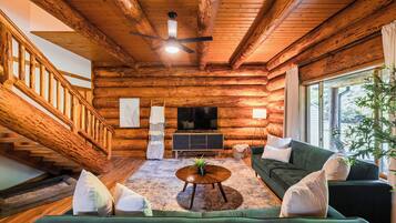 Interior de una cabaña de troncos con una escalera, un televisor, un sofá verde y una mesa de centro.