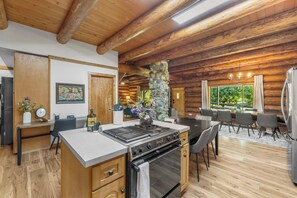 A modern kitchen with a central island, a dining area with a view, and a stone fireplace. - American Alps Lodge: Riverfront, Beach, Hot Tub, EV Charger, Game Room (Marblemount)