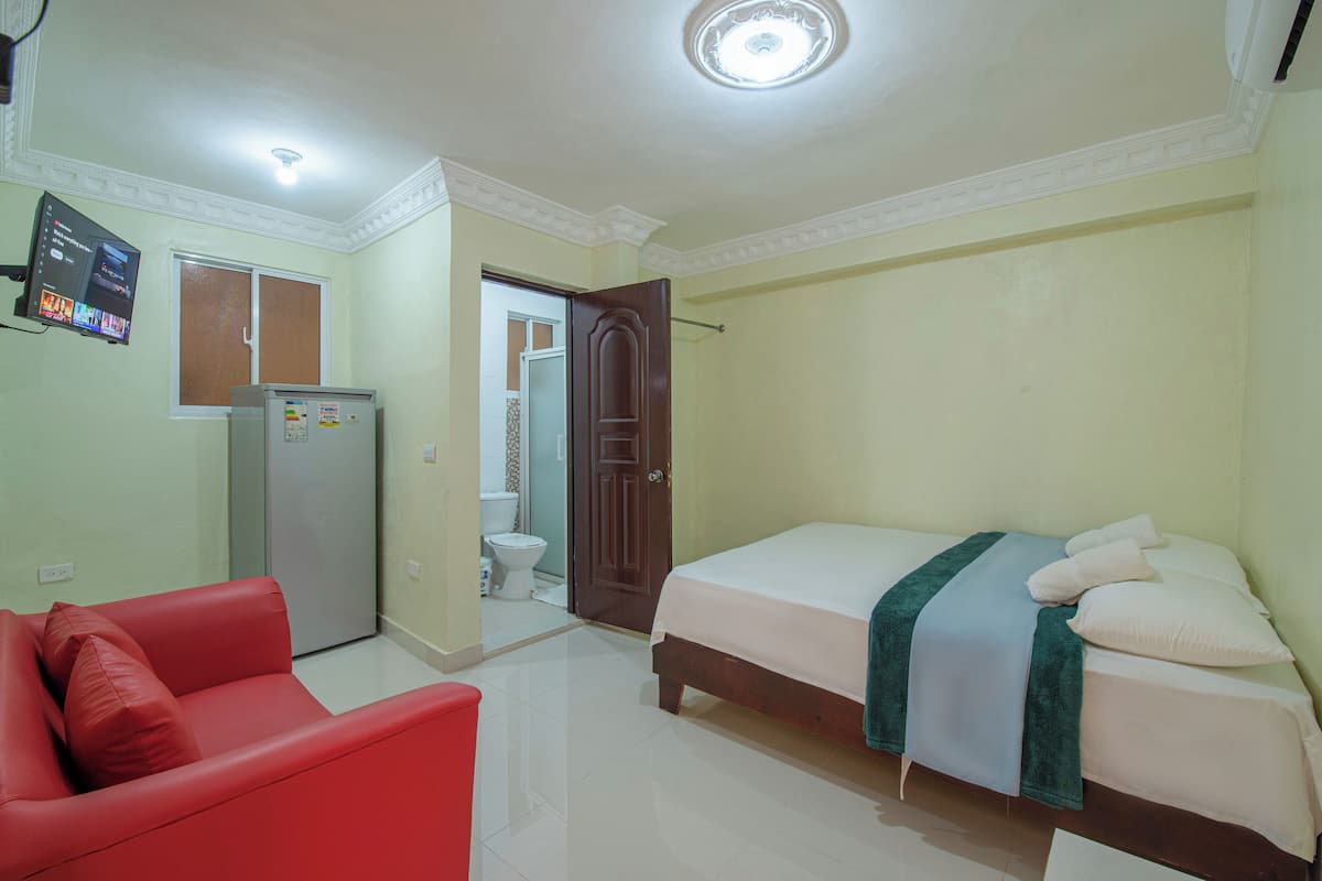 A bedroom with a bed, a red sofa, a refrigerator, and a toilet visible through an open door.