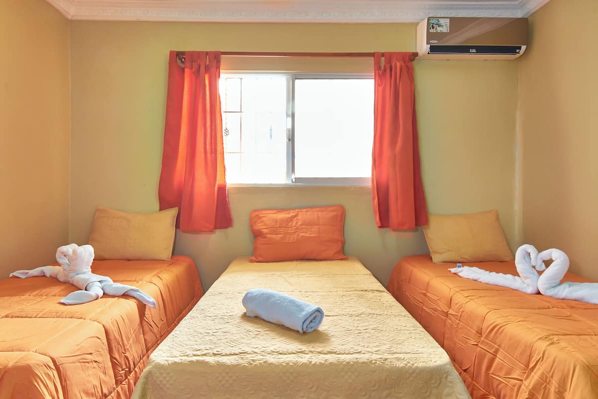 A hotel room with two beds, orange bedding, a central bed with a rolled towel, and a window with orange curtains.