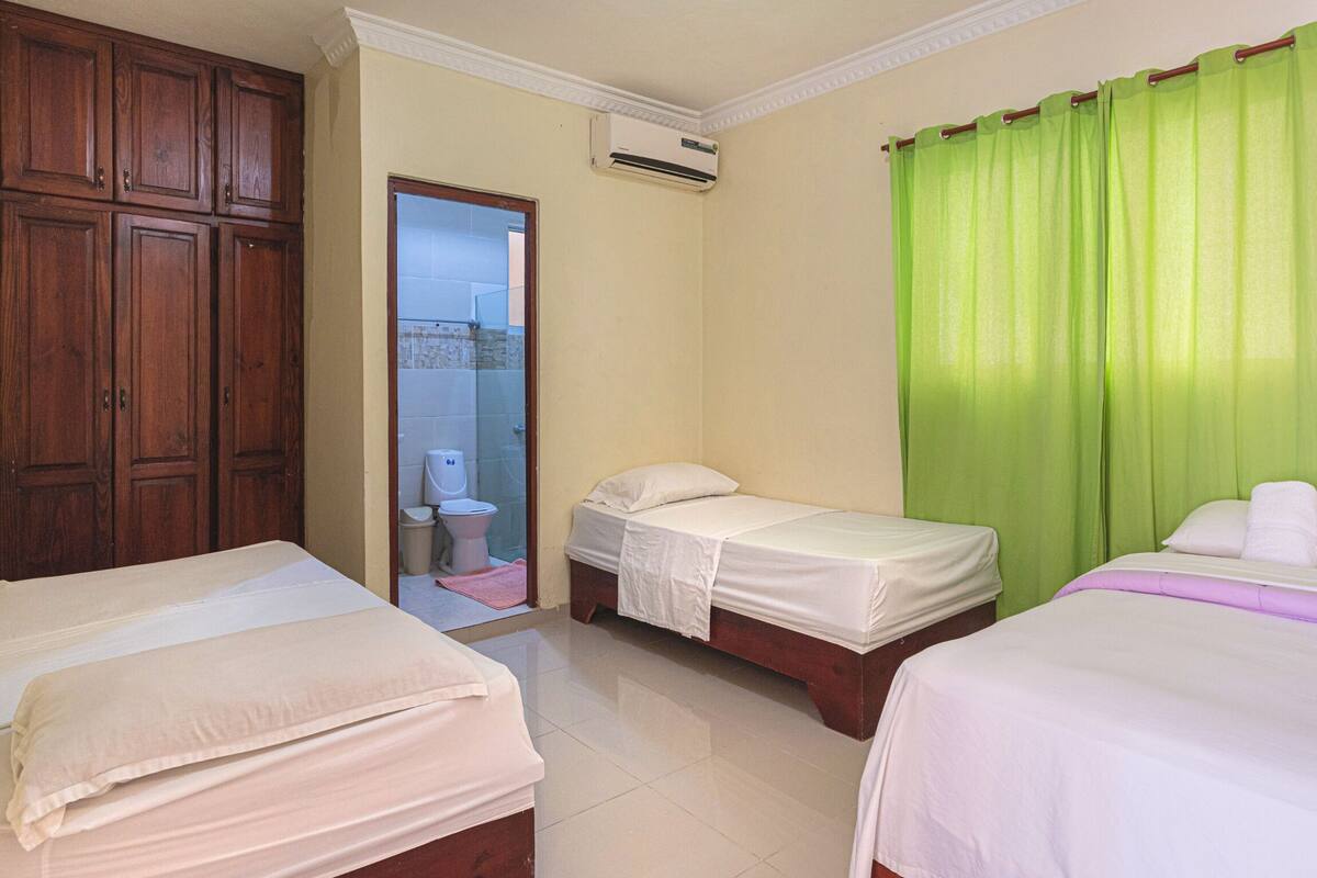 A hotel room with two beds, a wooden wardrobe, a bathroom with a toilet and shower, and a green curtain.