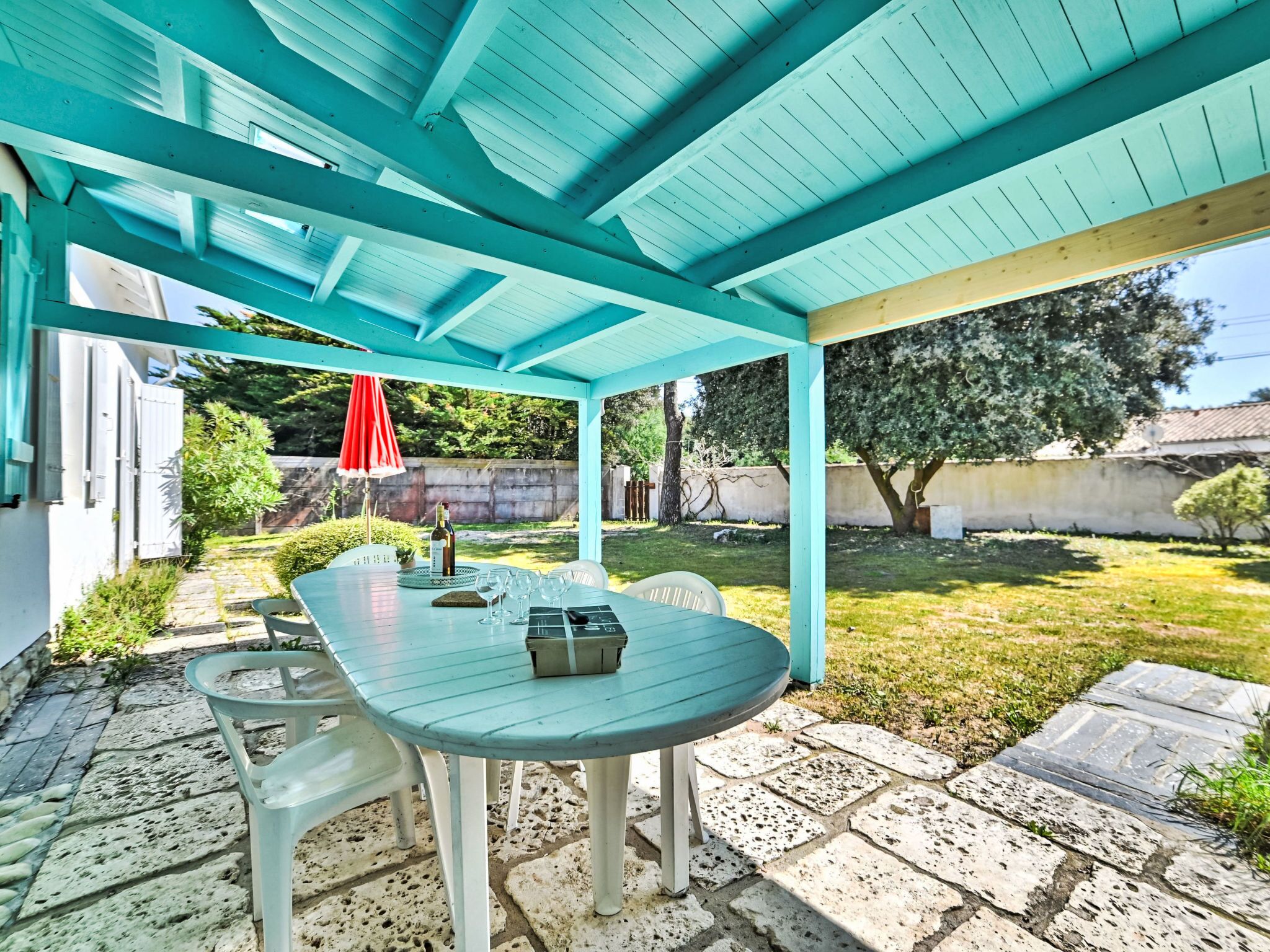 Un patio abrité avec une table et des chaises, un parasol rouge et une bouteille de vin.