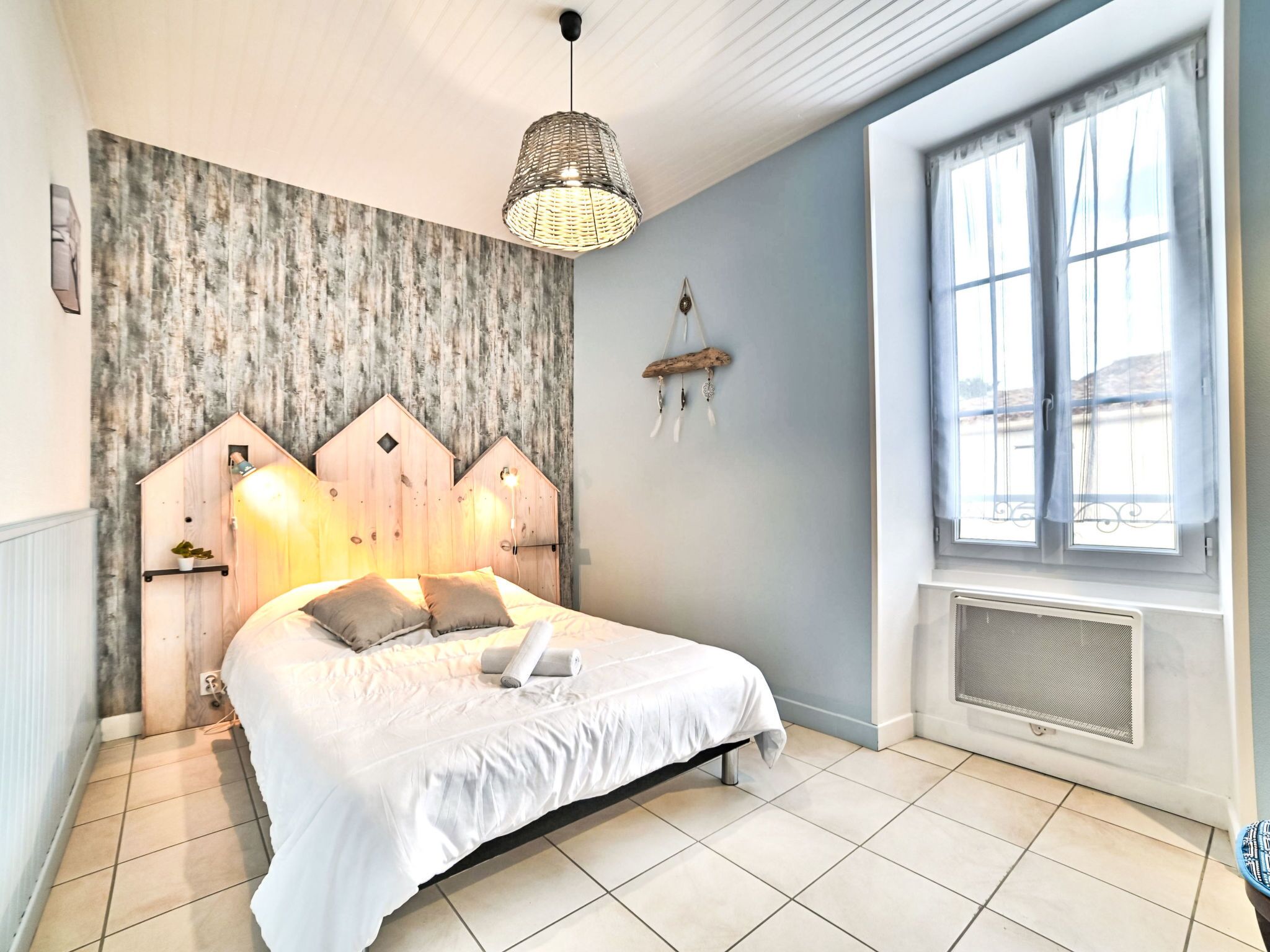 A bedroom with a bed, a wooden headboard, a hanging light fixture, a window with curtains, and a radiator.