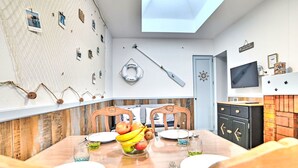 A dining area with a wooden table, chairs, and a fruit bowl. There is a ceiling light, a wall-mounted TV, and a door with a nautical-themed decoration. - Cheray 1 by Interhome (Ile d'Oléron)