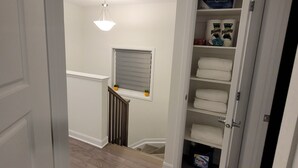 A hallway with a white door, a wooden floor, a ceiling light, a window with blinds, and a storage unit with towels and toilet paper.